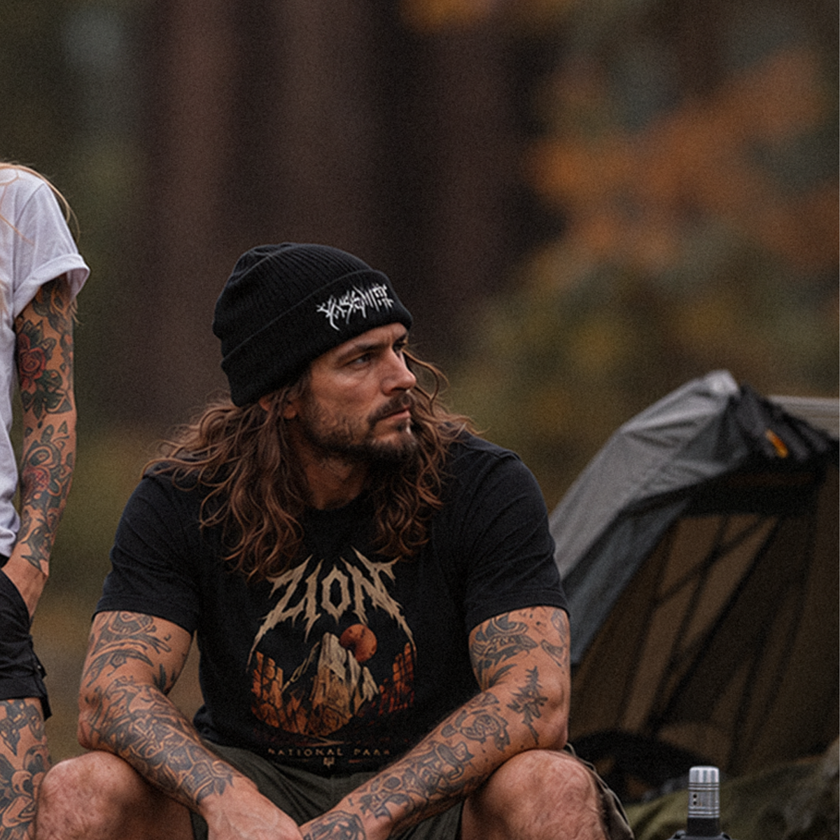 A man with long hair and tattoos sits outdoors wearing a black beanie with a white logo and a black graphic t-shirt. A tent is visible in the blurred background, along with another person partially cropped on the left.